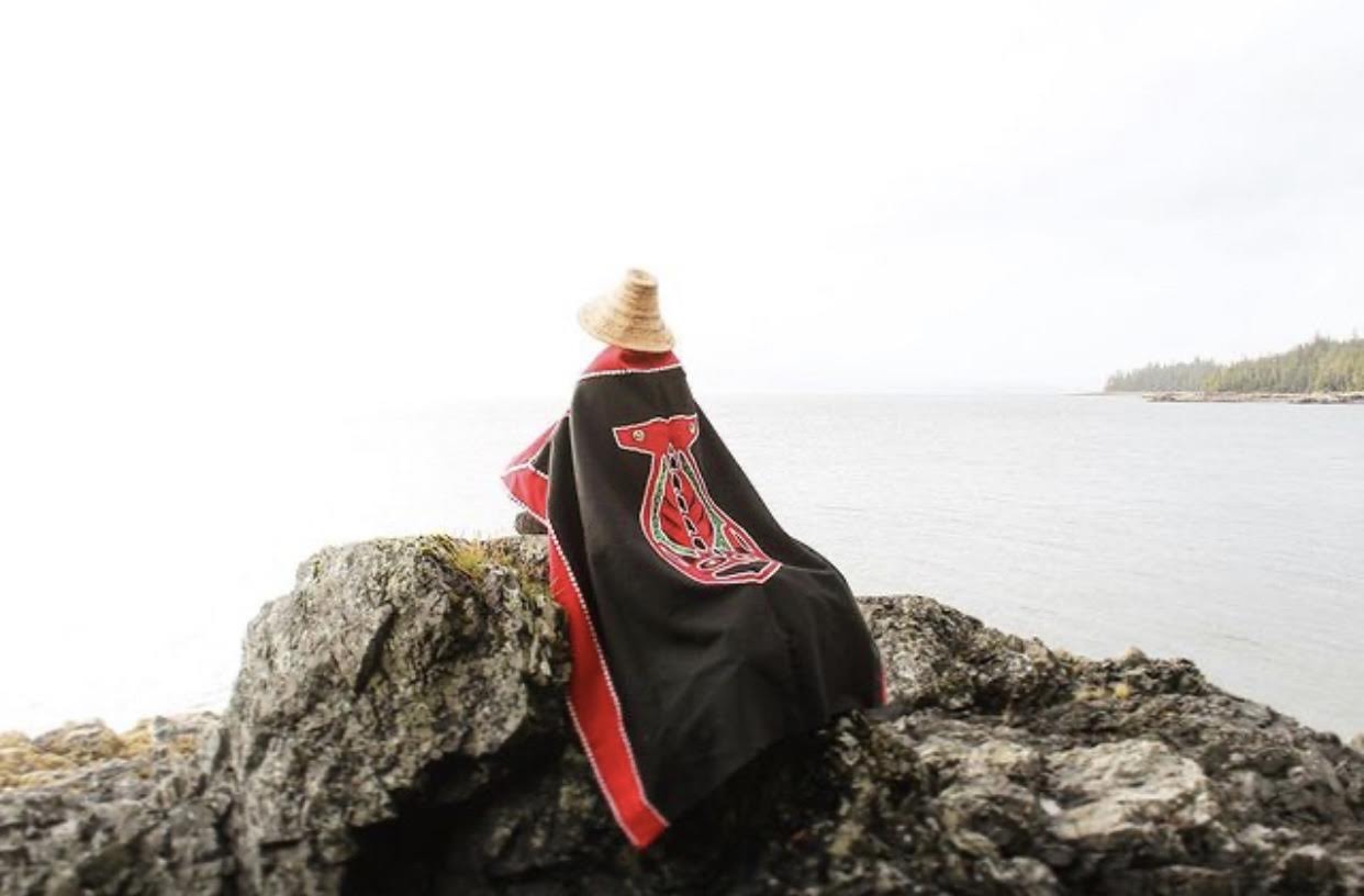Marina Anderson (Haida and Tlingt) in a traditional woven cedar hat and formline button blanket. Image taken with a self-timer on Tlingit and Haida shared lands. [Photo] Marina Anderson